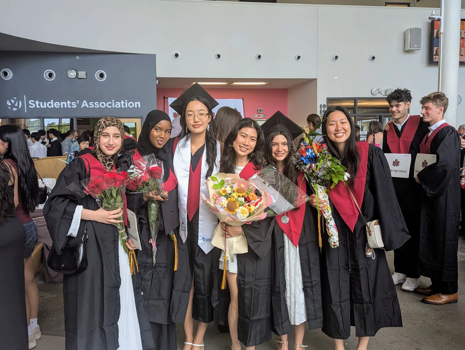 University graduates in academic gowns holding bouquets at a students' association convocation reception.