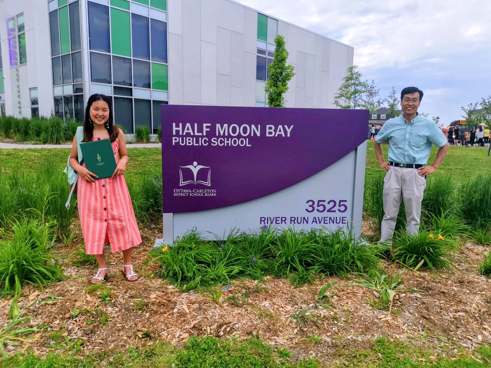 A child and an adult standing on either side of the Half Moon Bay Public School sign on River Run Avenue.