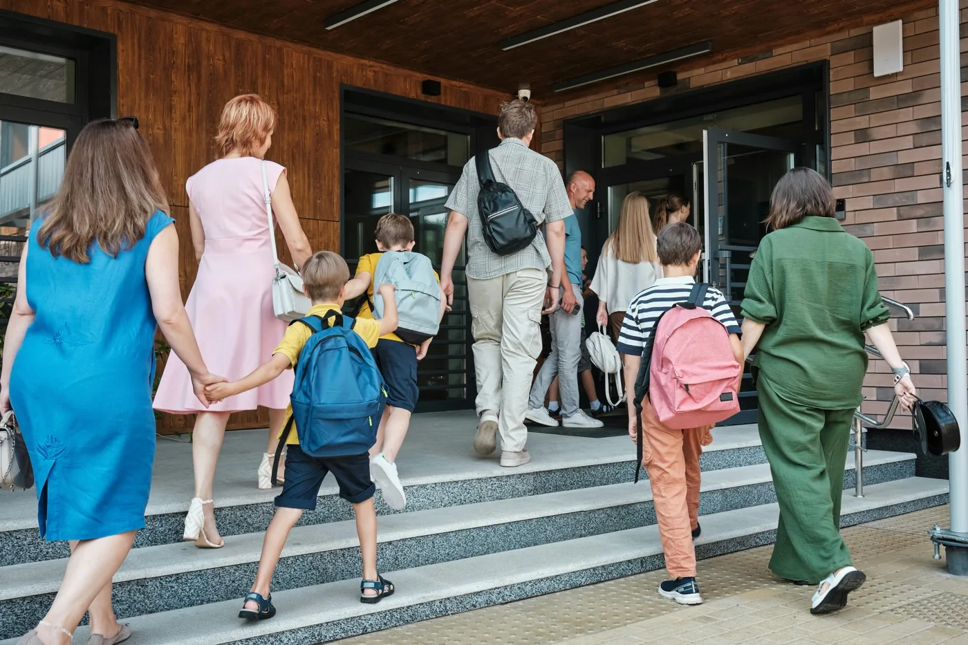 Families and children with backpacks climbing the steps into a school entrance