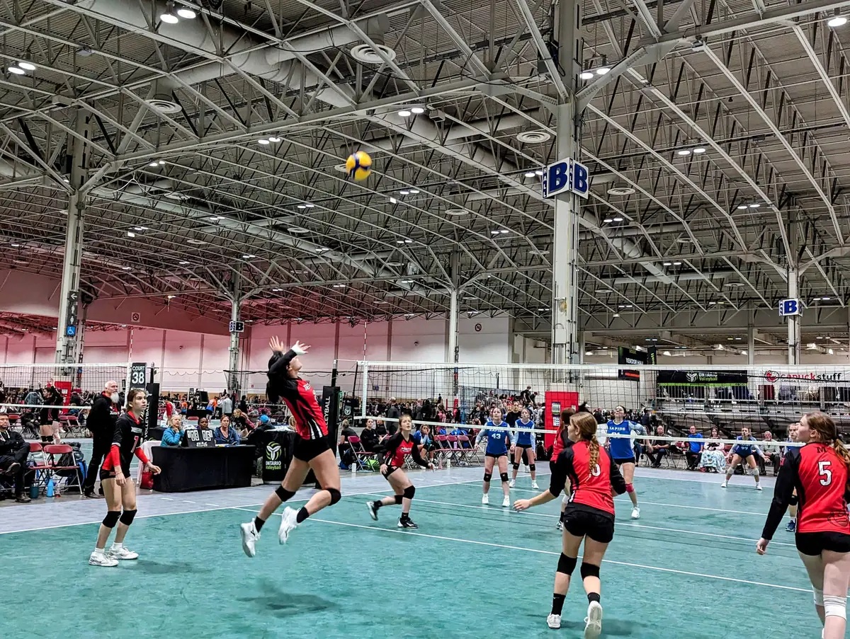 Young athletes mid-rally at an indoor volleyball tournament in a fieldhouse.